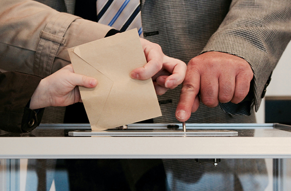 A close-up photograph of several people placing an envelope into a ballot box.